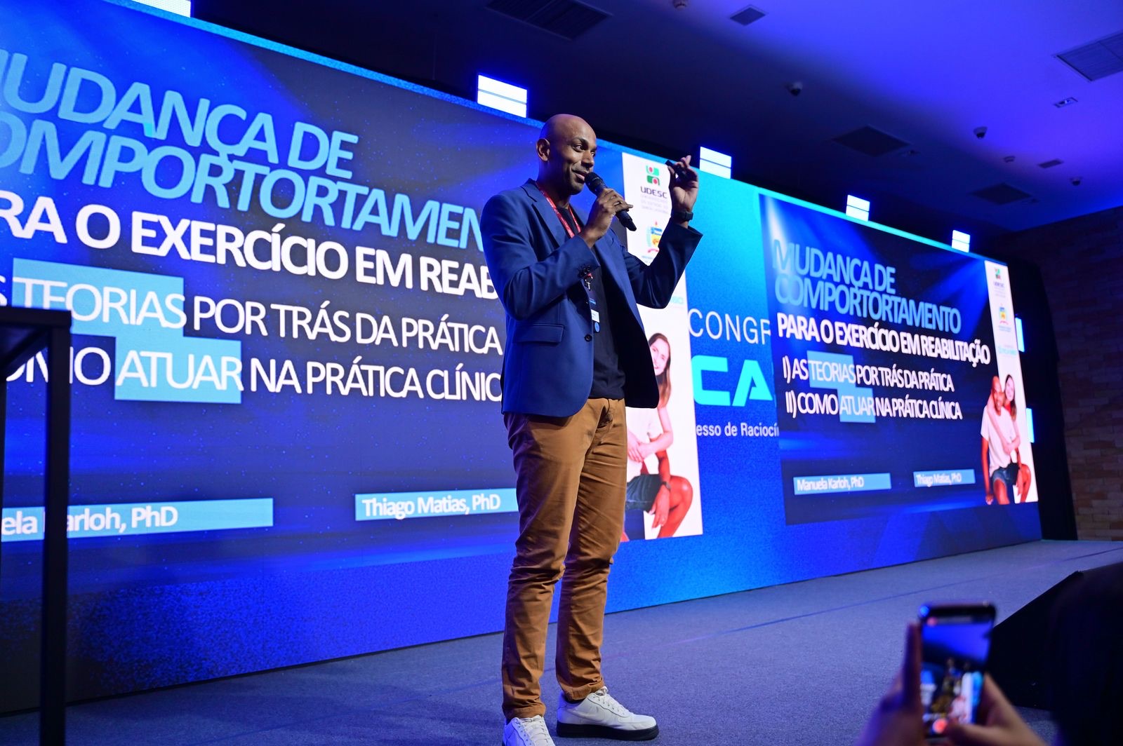 Thiago Matias palestrando sobre Mudança de Comportamento para o Exercício em Reabilitação, ao lado de telão com Manuela Karloh